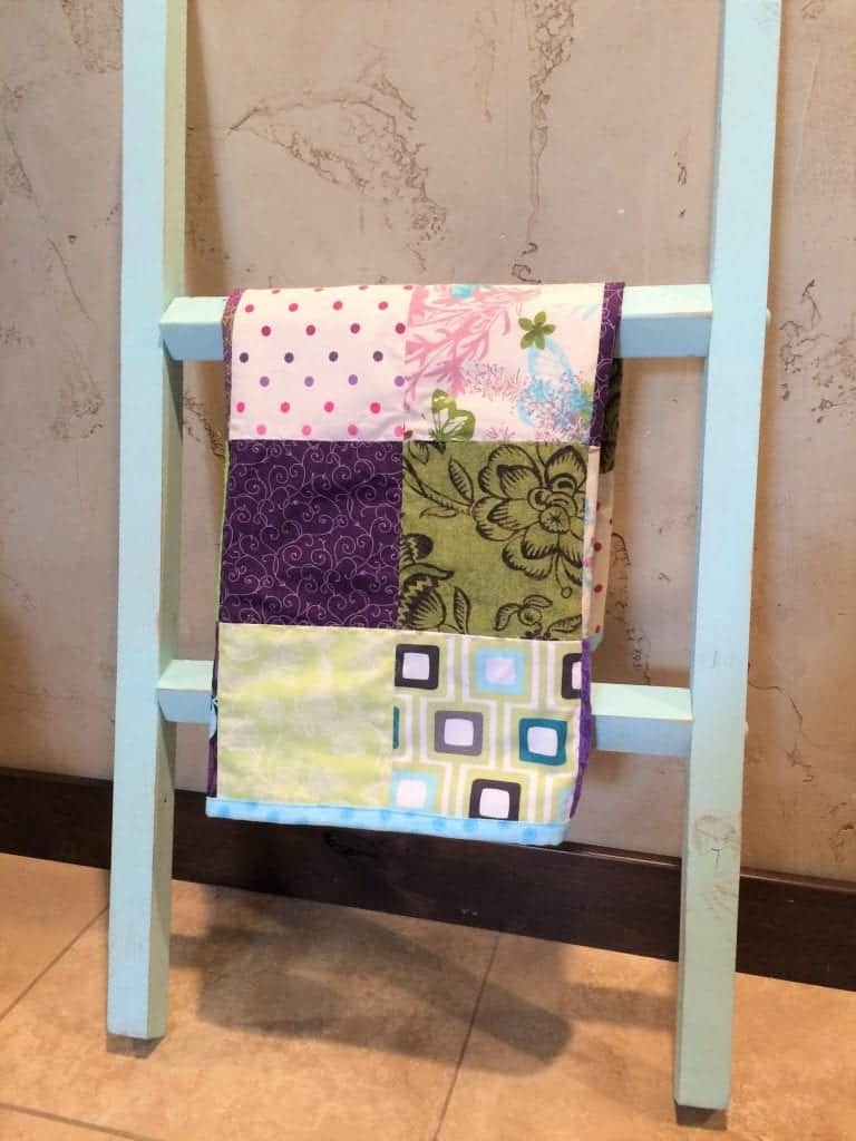 Baby quilt in purples and greens hanging from blue ladder.