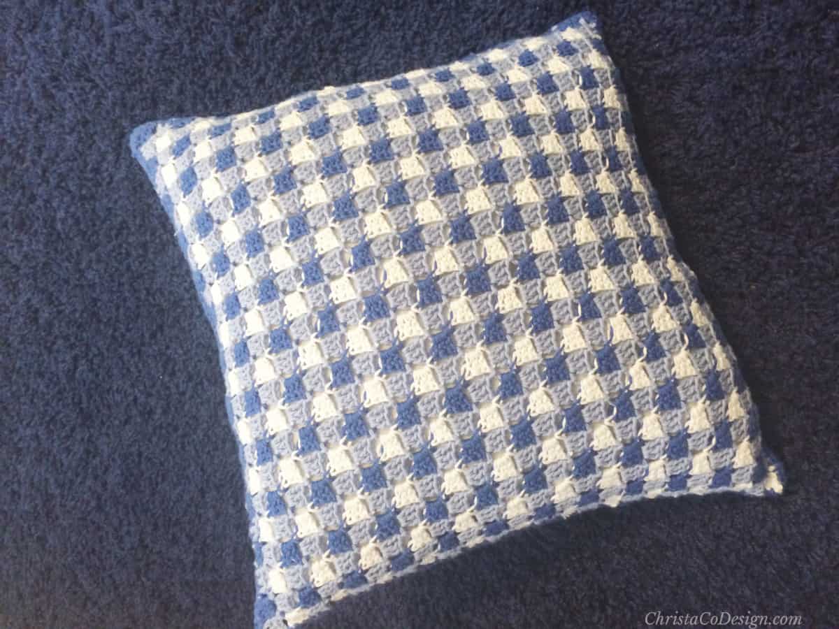 Blue and white plaid crochet pillow on dark carpet.