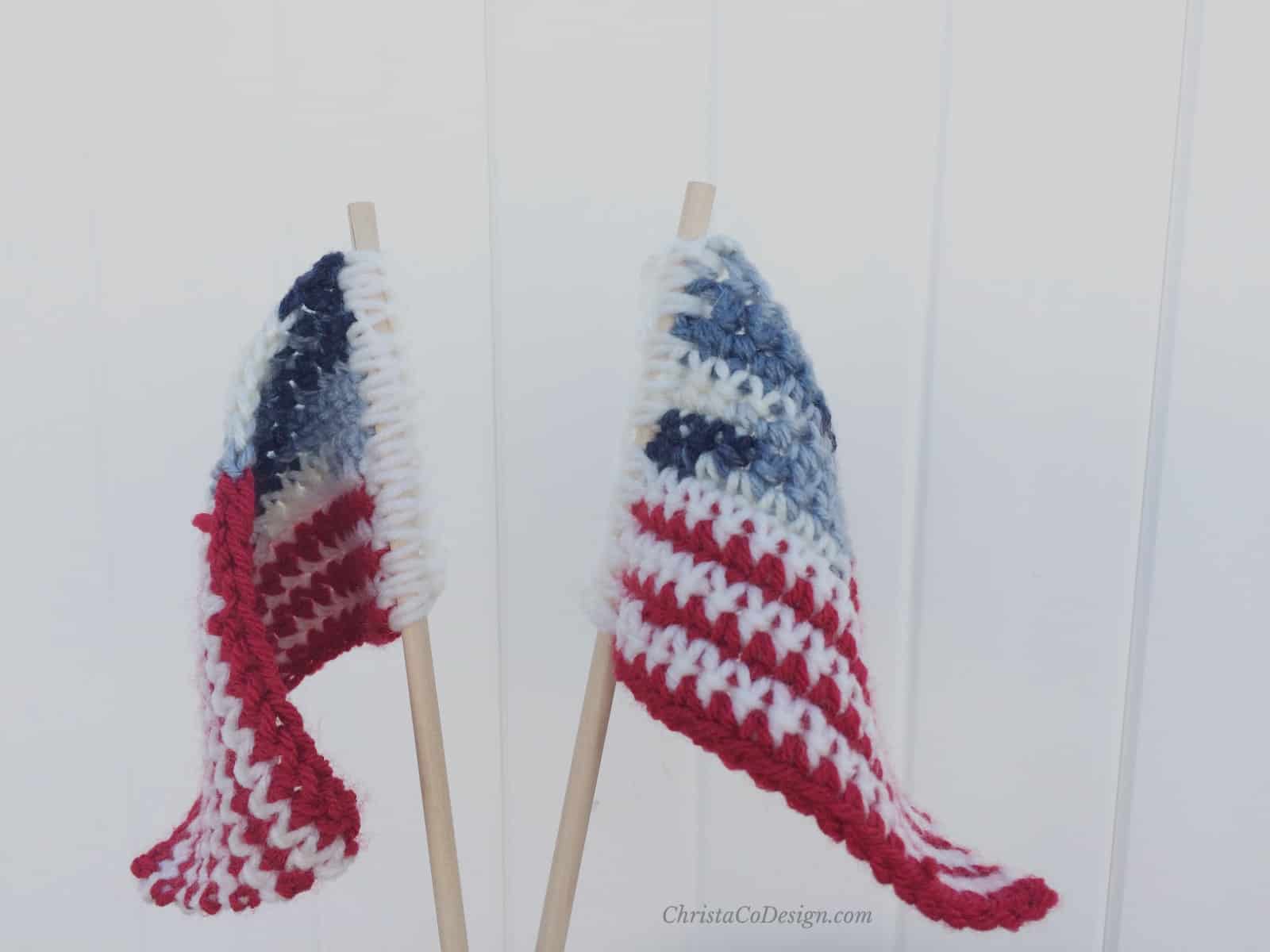 Two small crochet American flags on dowels in front of white fence.  