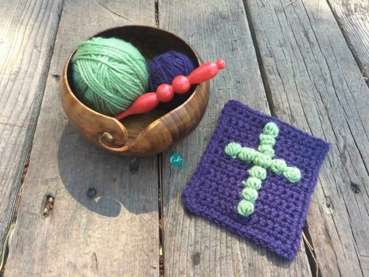 Purple prayer cloth with crochet cross in green on deck with yarn bowl and hook.