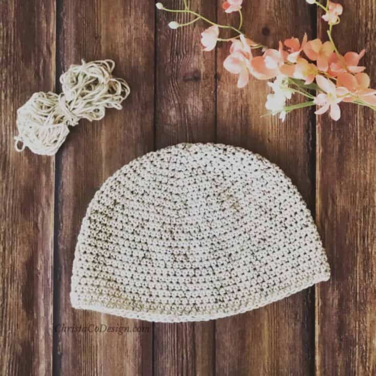 Chemo cap in cream with green flecks next to yarn and pink flowers.