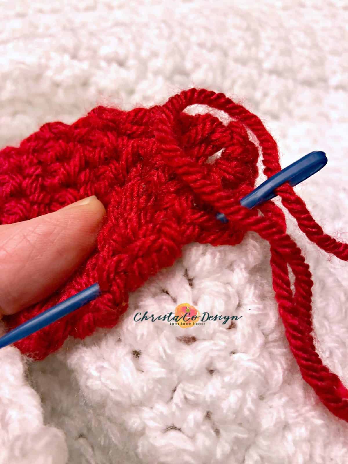 picture of blue yarn needle sewing through red crochet christmas light on to white blanket