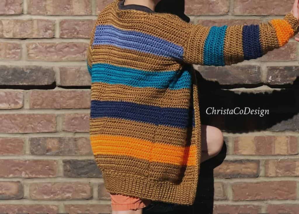 Boy reaching out in striped brown, orange and blue crochet cardigan in front of brick wall.