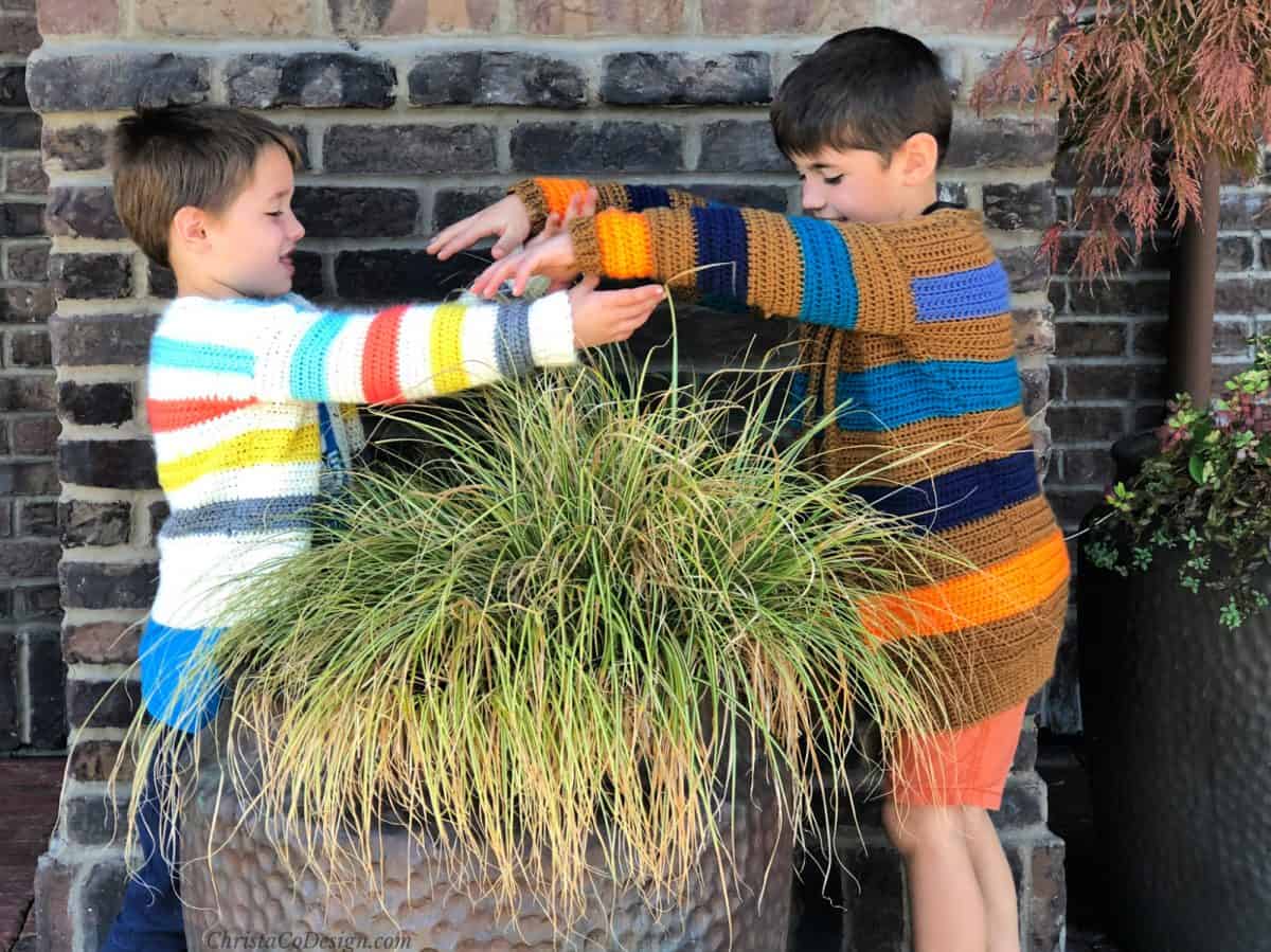 Two boys in their striped crochet cardigans reaching over plant in pot.