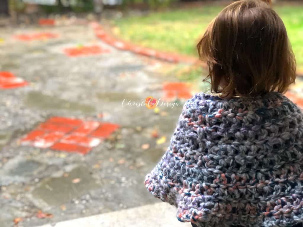 Toddler sitting in chunky crochet poncho and staring out at rain.