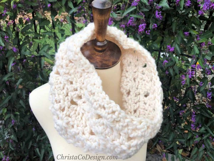 Chunky crochet neck warmer on mannequin in n front of greenery.