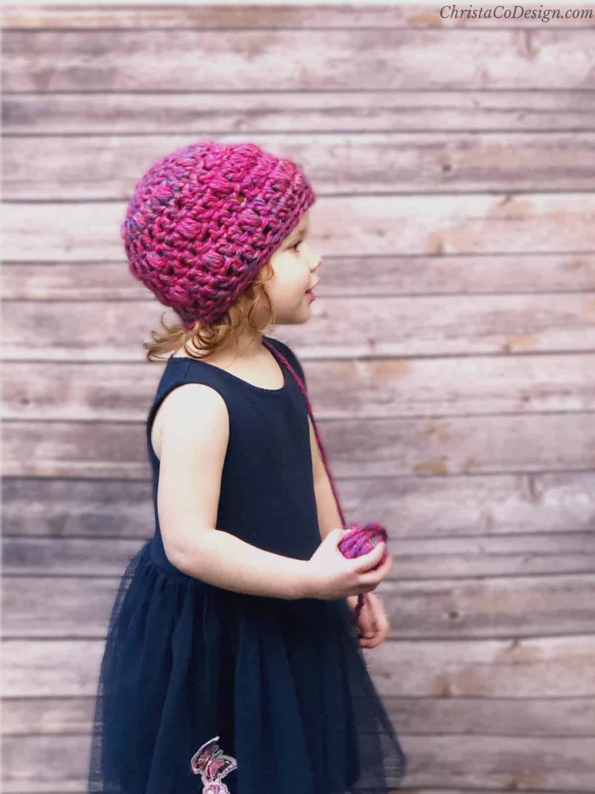 Girl in pink crochet beanie turned to the side with ball of yarn in hand.
