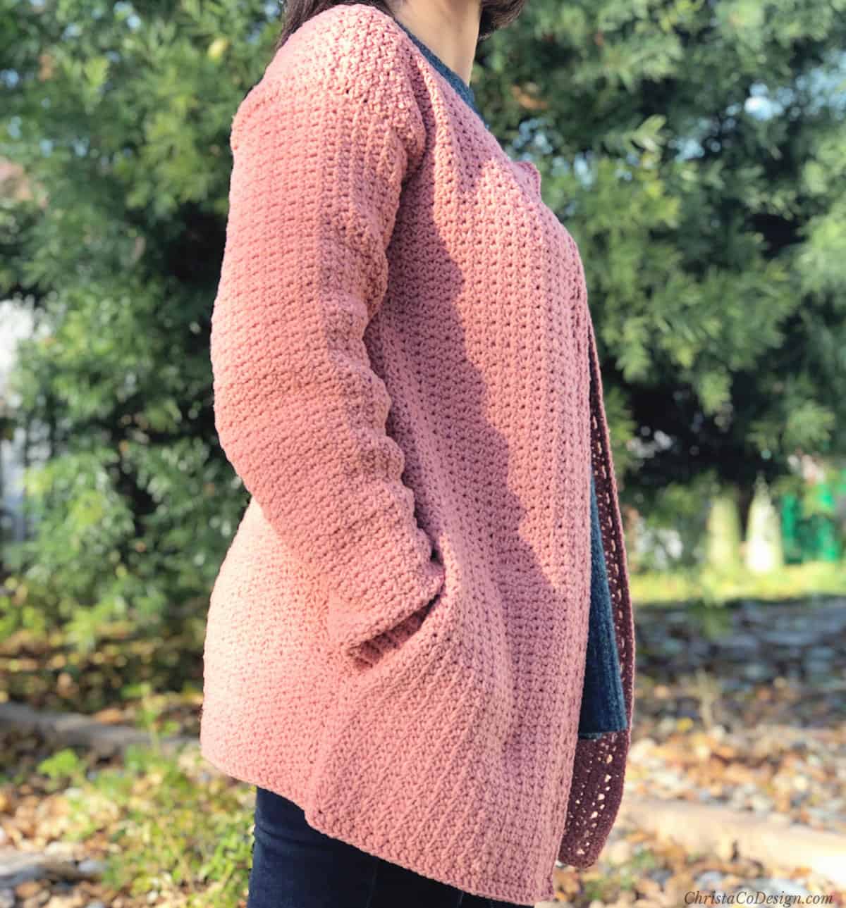 Woman with pink crochet cardigan on turned to side with hands in side seam pockets.