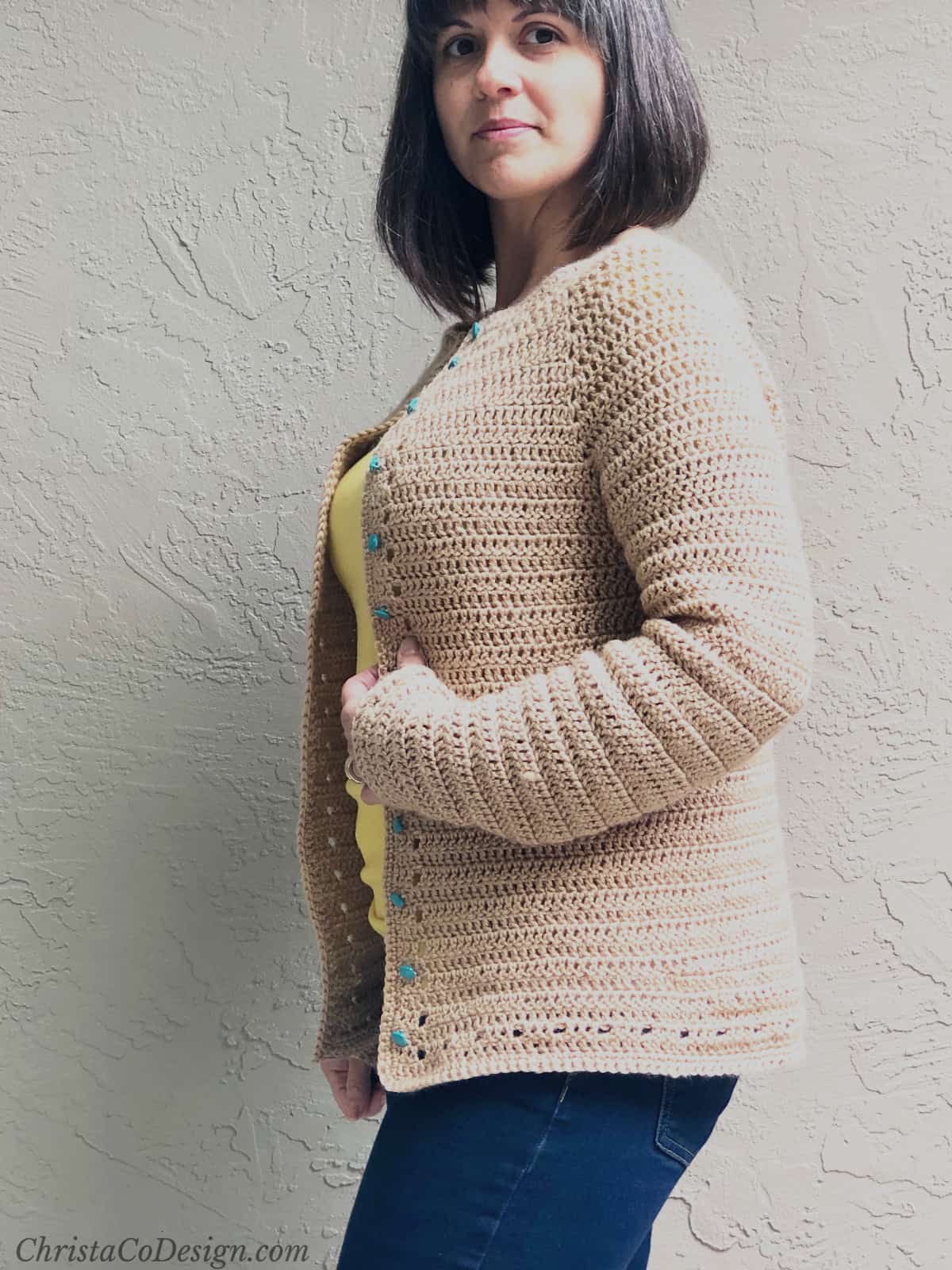 Woman turned to the side in beige crochet cardigan with elbow bent and smiling at camera.