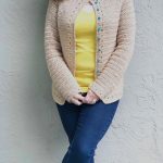 Woman in beige crochet cardigan leaning on wall in dark jeans.