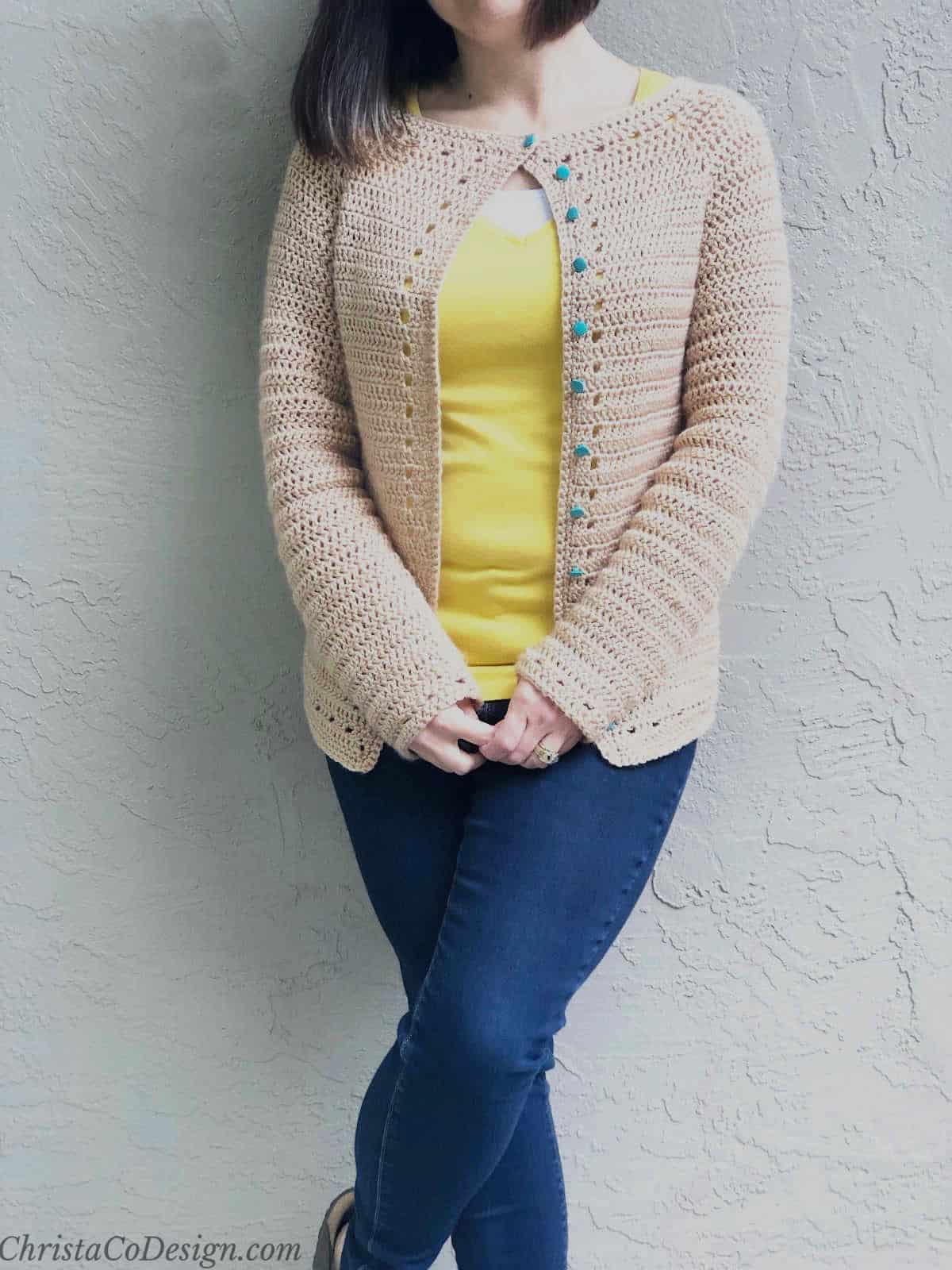 Woman leaning on wall with caramel colored crochet cardigan and dark jeans on crossed legs and holding hands together in front of her.