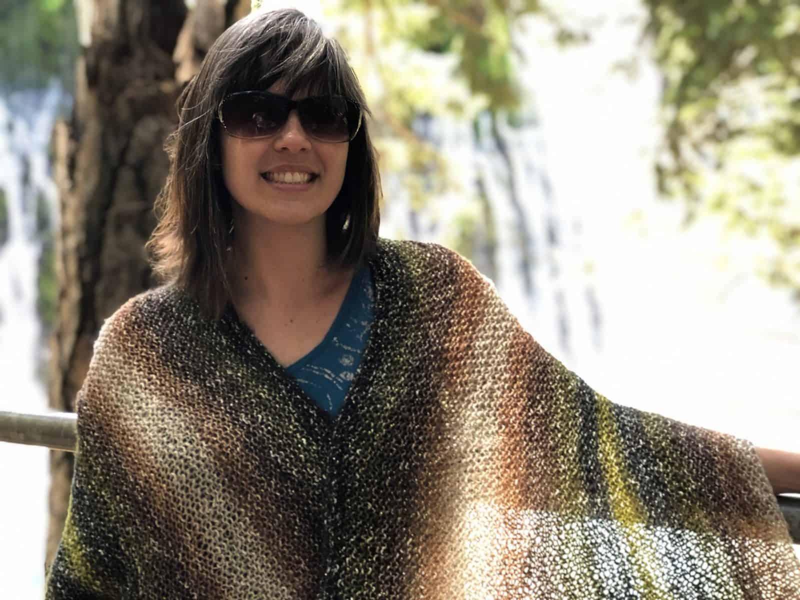 Woman in front of waterfall with light filtering through garter stitch knit shawl in greens and brown.