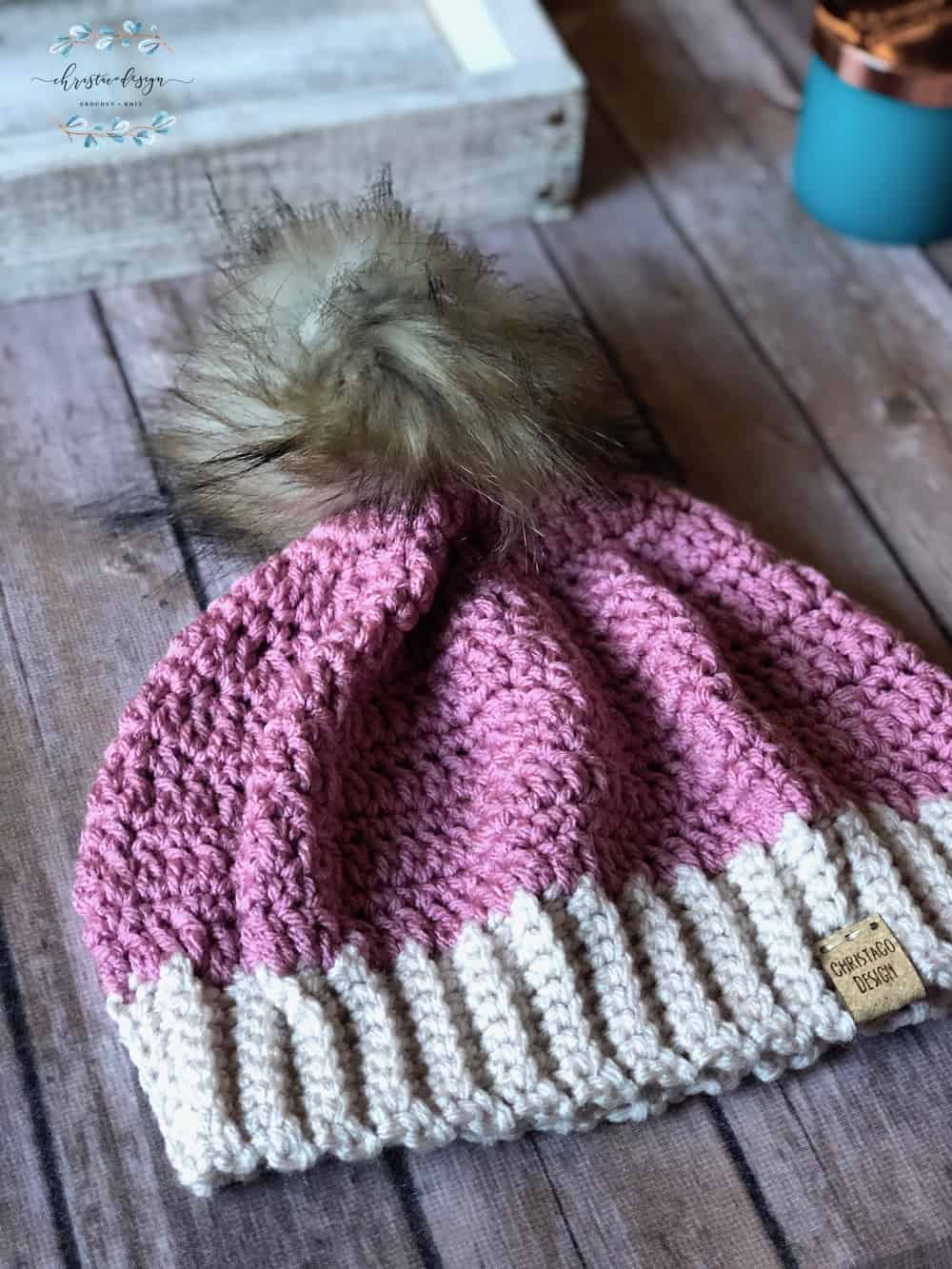 Pink crochet hat with fur pom on wood table.