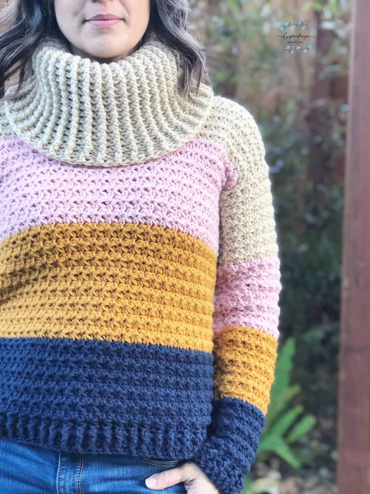 Woman in crochet cowl neck sweater of beige, pink, gold and navy color blocks.