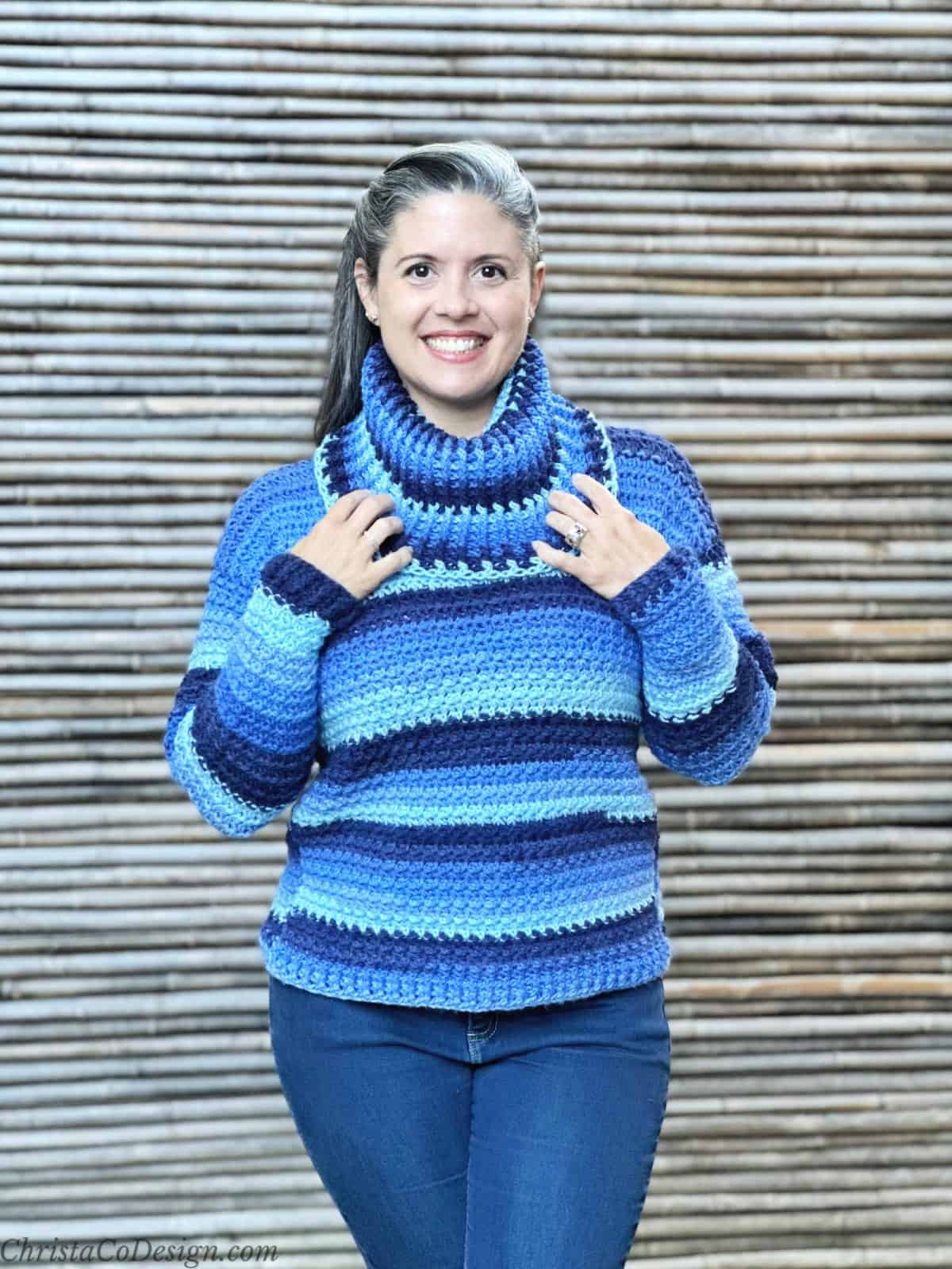 Woman in blue striped crochet sweater smiling with hands on cowl neck.