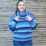 Woman in blue striped crochet sweater smiling with hands on cowl neck.