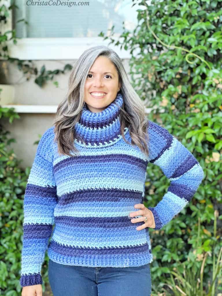Woman in long grey and brown hair smiling at camera in crochet sweater of blue stripes.