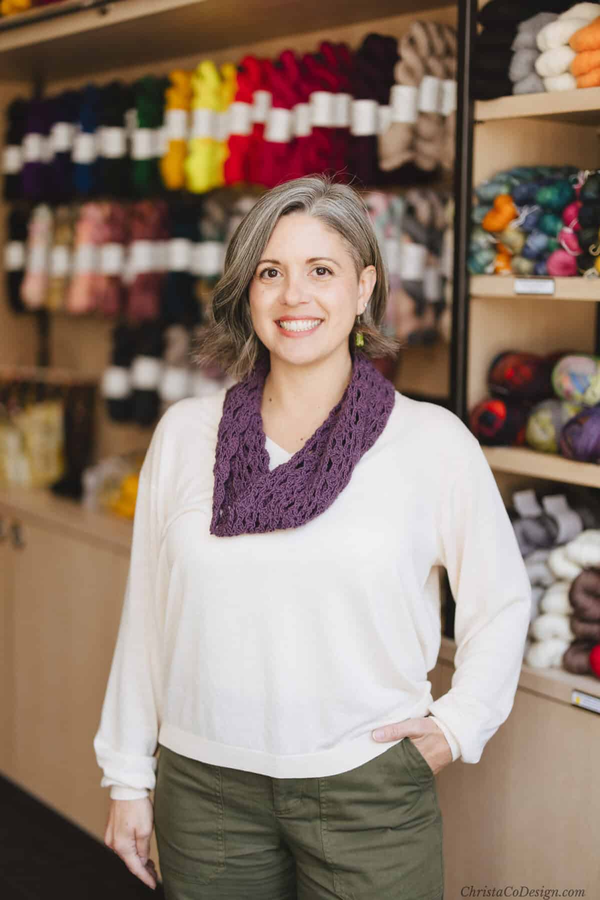 Woman with purple crochet cowl over white sweater in front of wall of yarn.