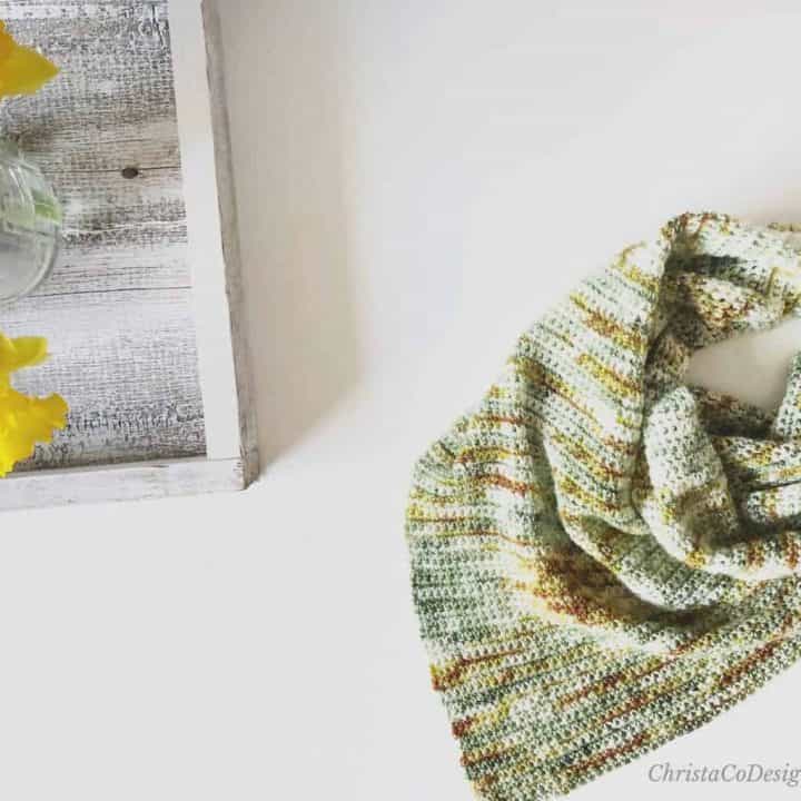 Crochet triangle scarf in hand dyed yarn on table with daisies.