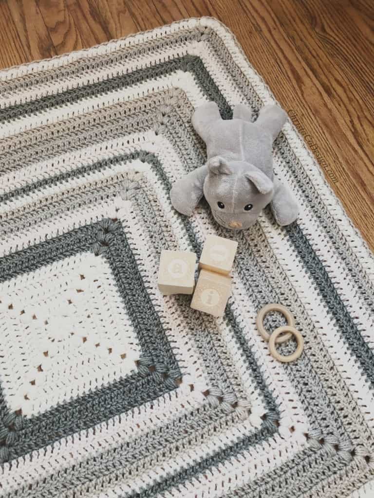 Grey and white striped square crochet blanket with wood blocks and stuff cat on side.