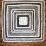 Center out crochet blanket in grey and white laid out flat on wood floor.
