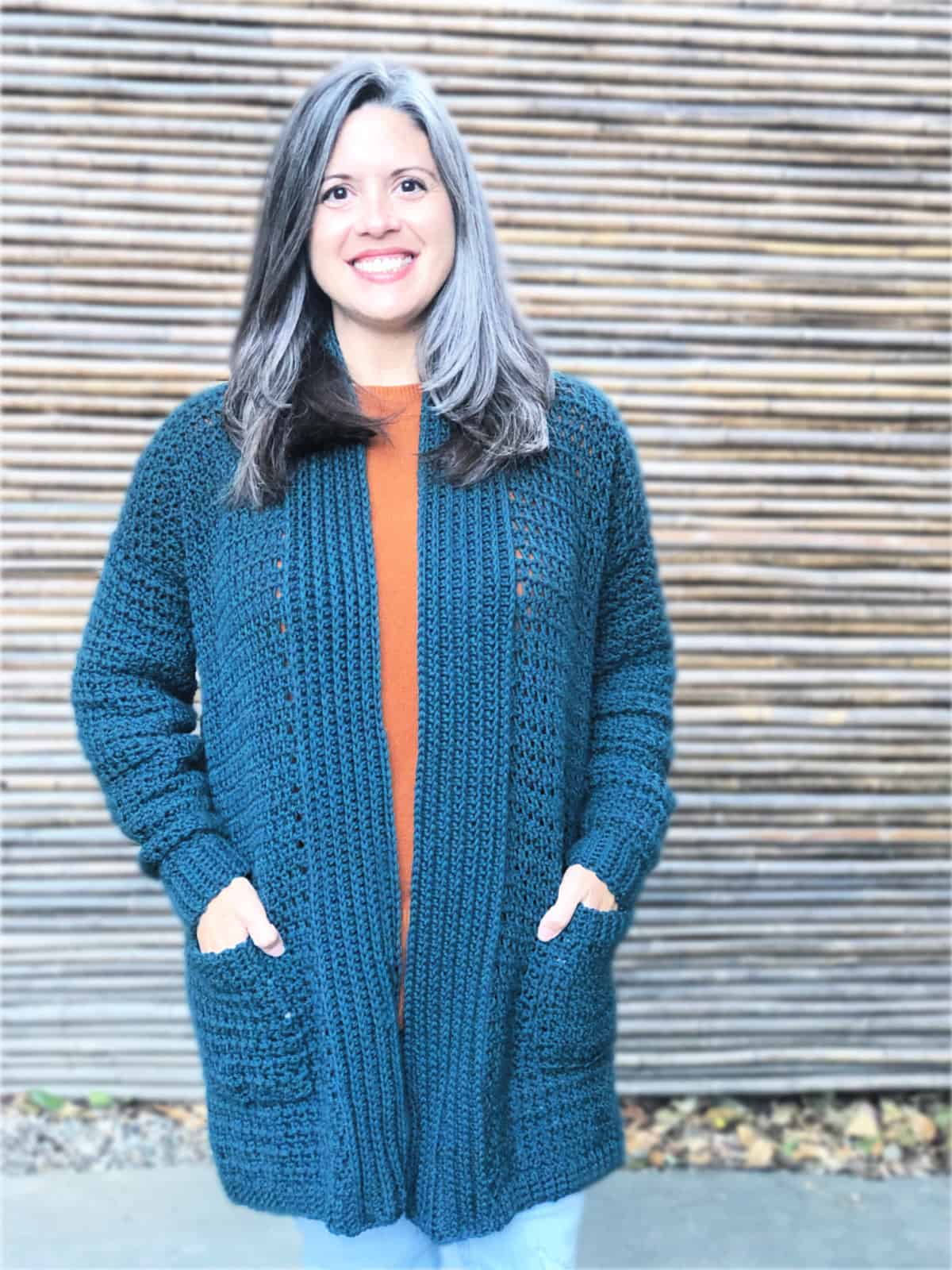 Woman smiling in with hands in pockets of blue green crochet cardigan.