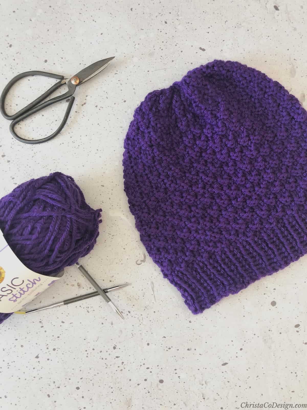 The purple knit hat is laid flat with yarn, needle and scissors.