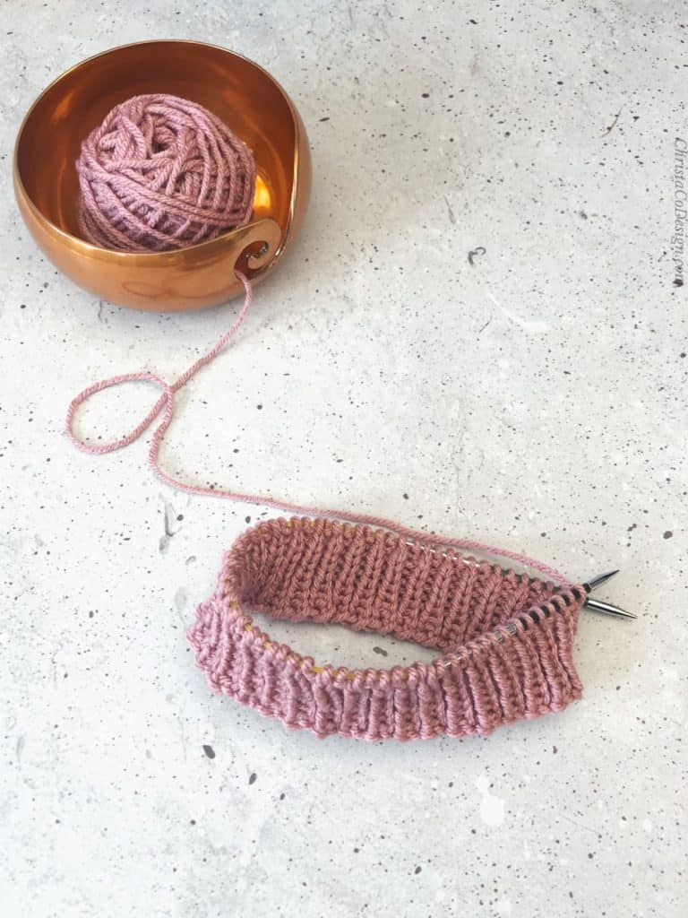 Pink ribbing on needles with yarn in bowl.