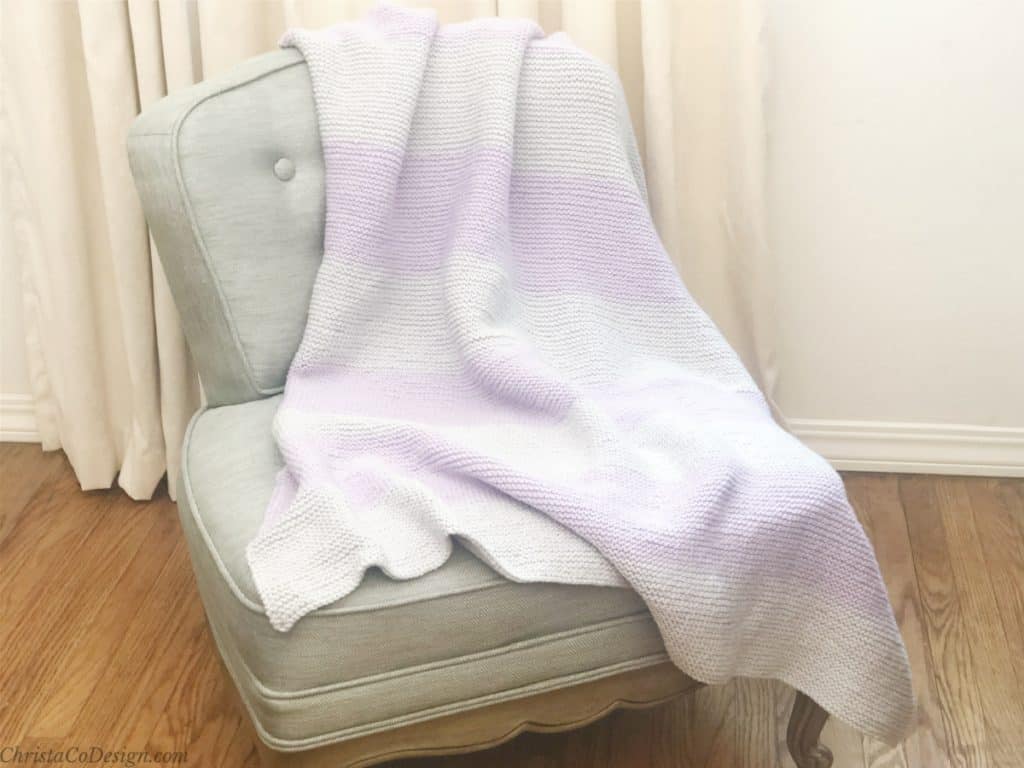 Purple and silver knitted blanket draped on chair in front of cream curtain.