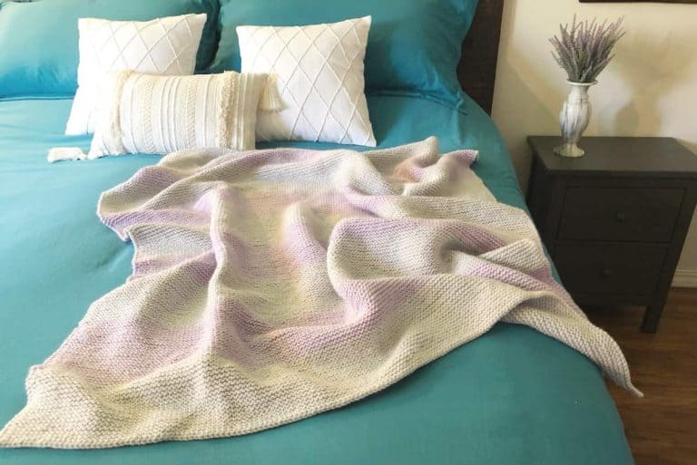 Striped purple and grey knit blanket on blue covered bed with white pillows.