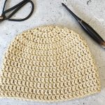 Top down double crochet beanie in cream yarn with hook laid flat.