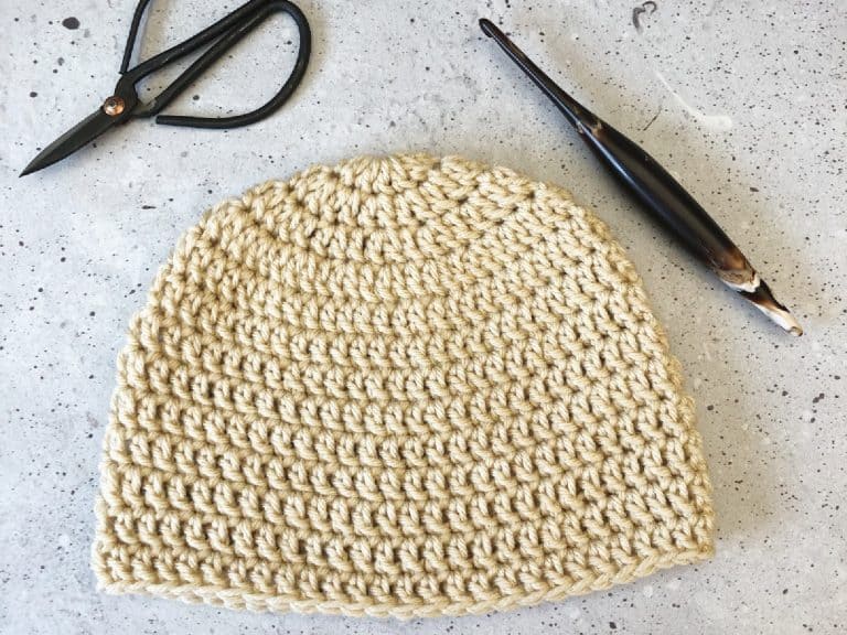Top down double crochet beanie in cream yarn with hook laid flat.