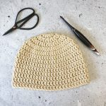 Double crochet beanie in beige yarn, scissors, crochet hook on grey speckled background.