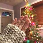 Chunky fingerless gloves on woman's hand in front of lit Christmas tree covered in colorful lights.