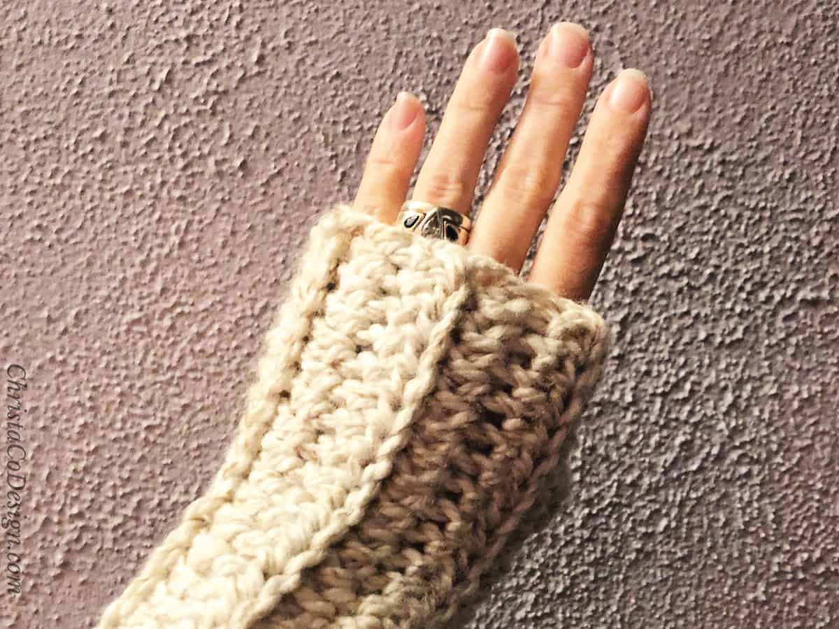Woman's hand wrapped in cozy chunky fingerless glove in front of lavender wall.