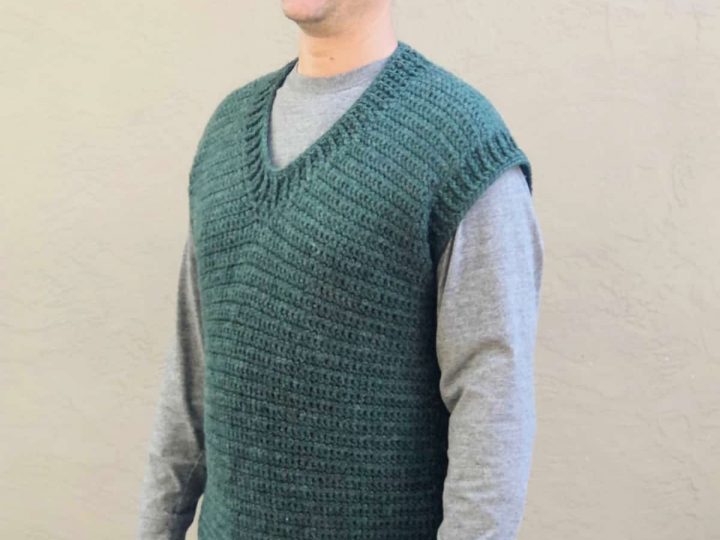 Man smiling while wearing green crochet v-neck sweater vest in front of beige wall.