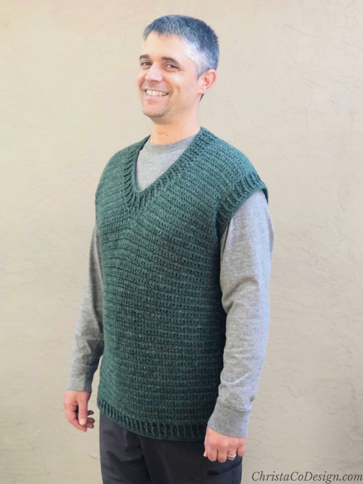 Man smiling while wearing green crochet v-neck sweater vest in front of beige wall.