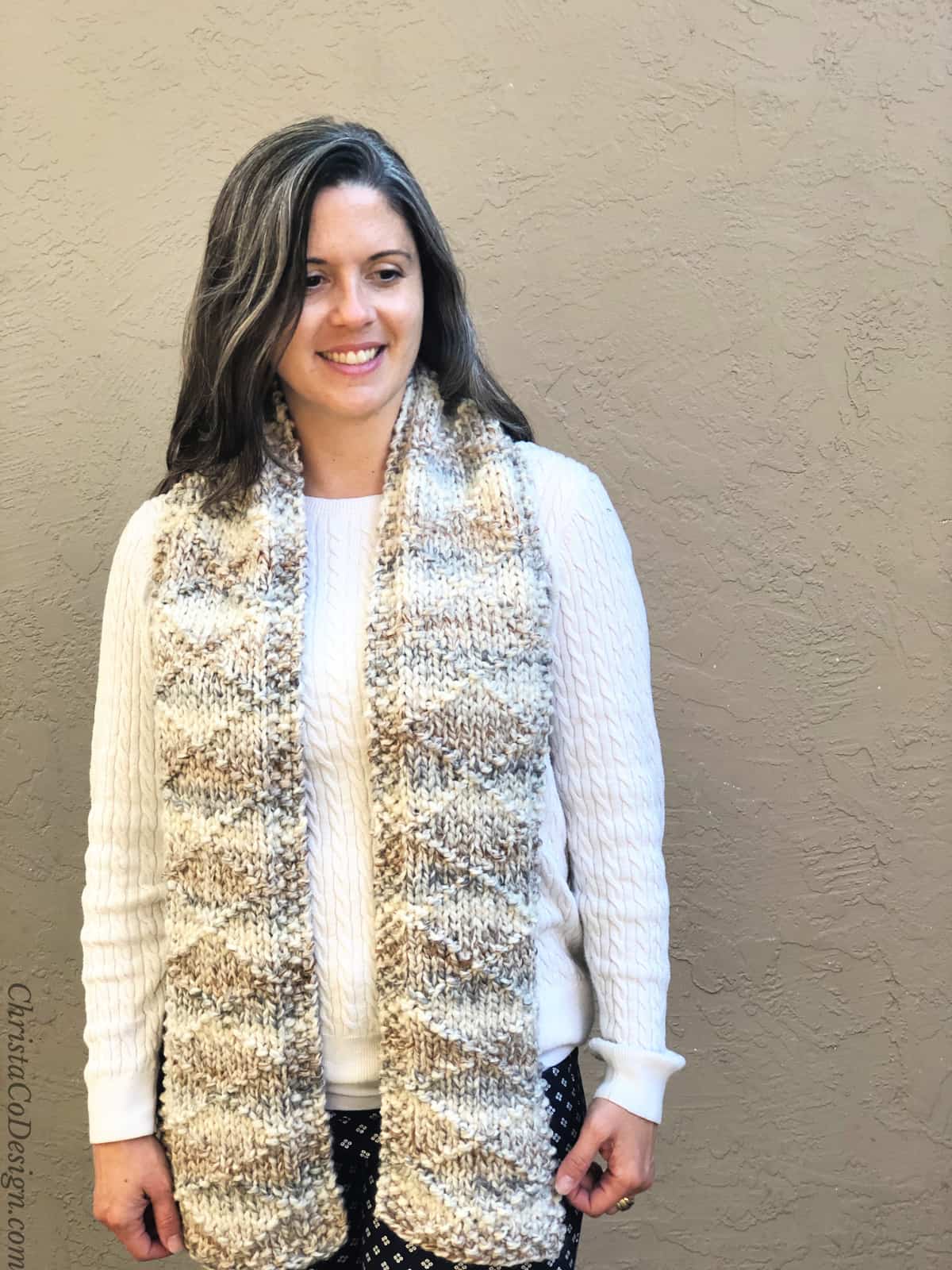 Woman in chunky knit scarf in beiges over white sweater smiling and looking down.