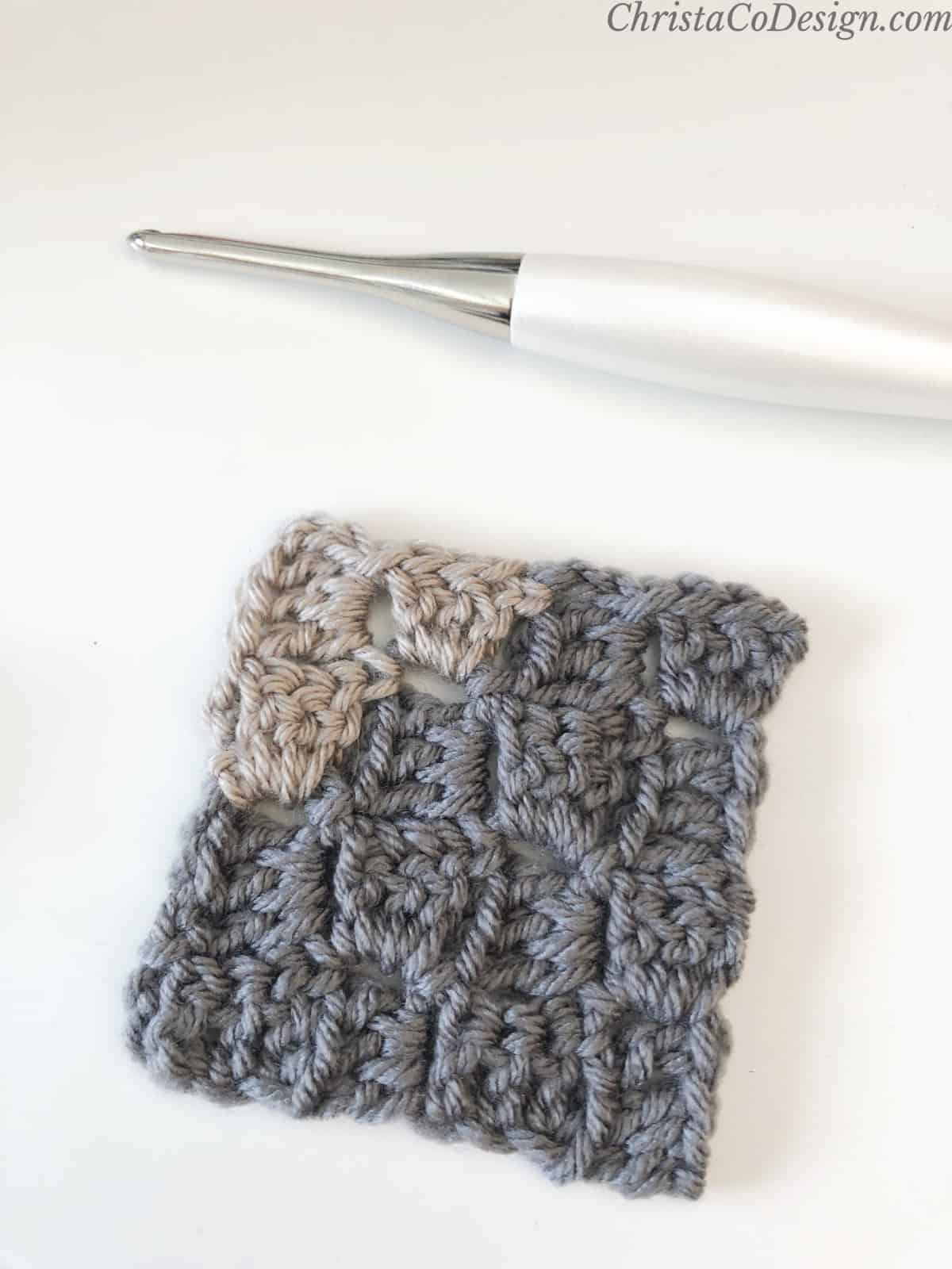 Grey crochet corner to corner square with beige color change at one corner on white table with crochet hook.