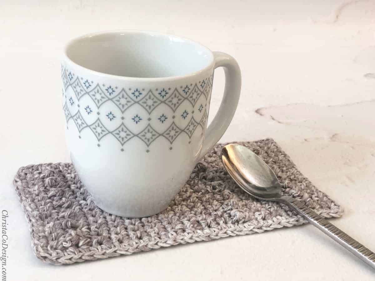 White coffee mug on grey crochet mug rug with spoon also.