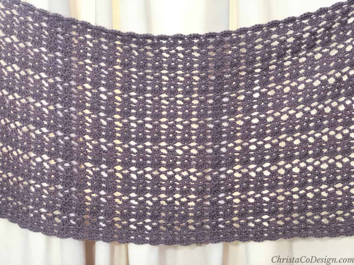 Lacy purple rectangle crochet shawl pinned to full width on cream curtain.