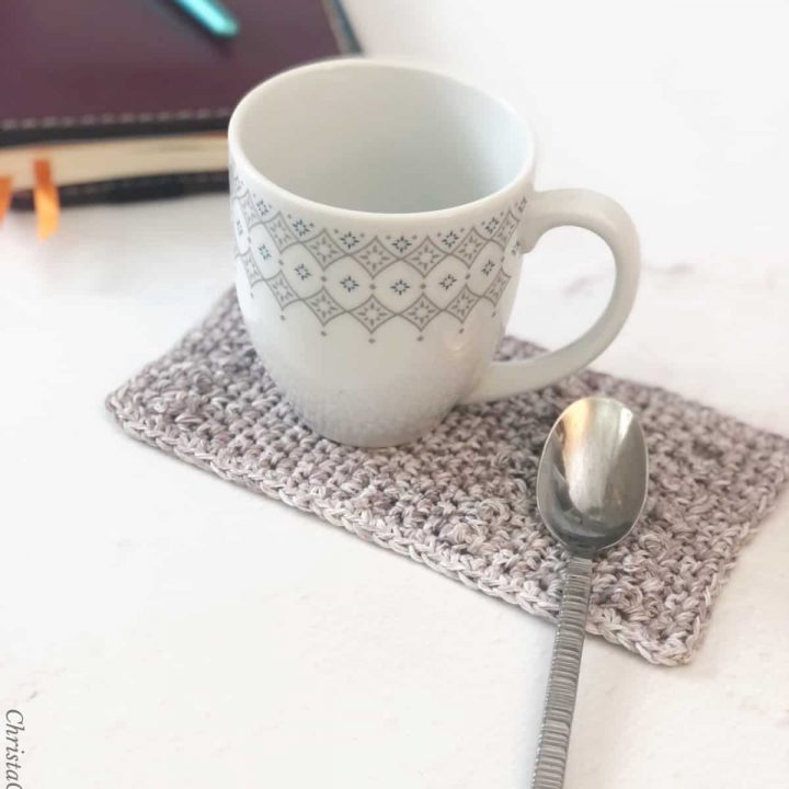 Coffee mug on grey crochet mug rug with spoon area and notebook on table.