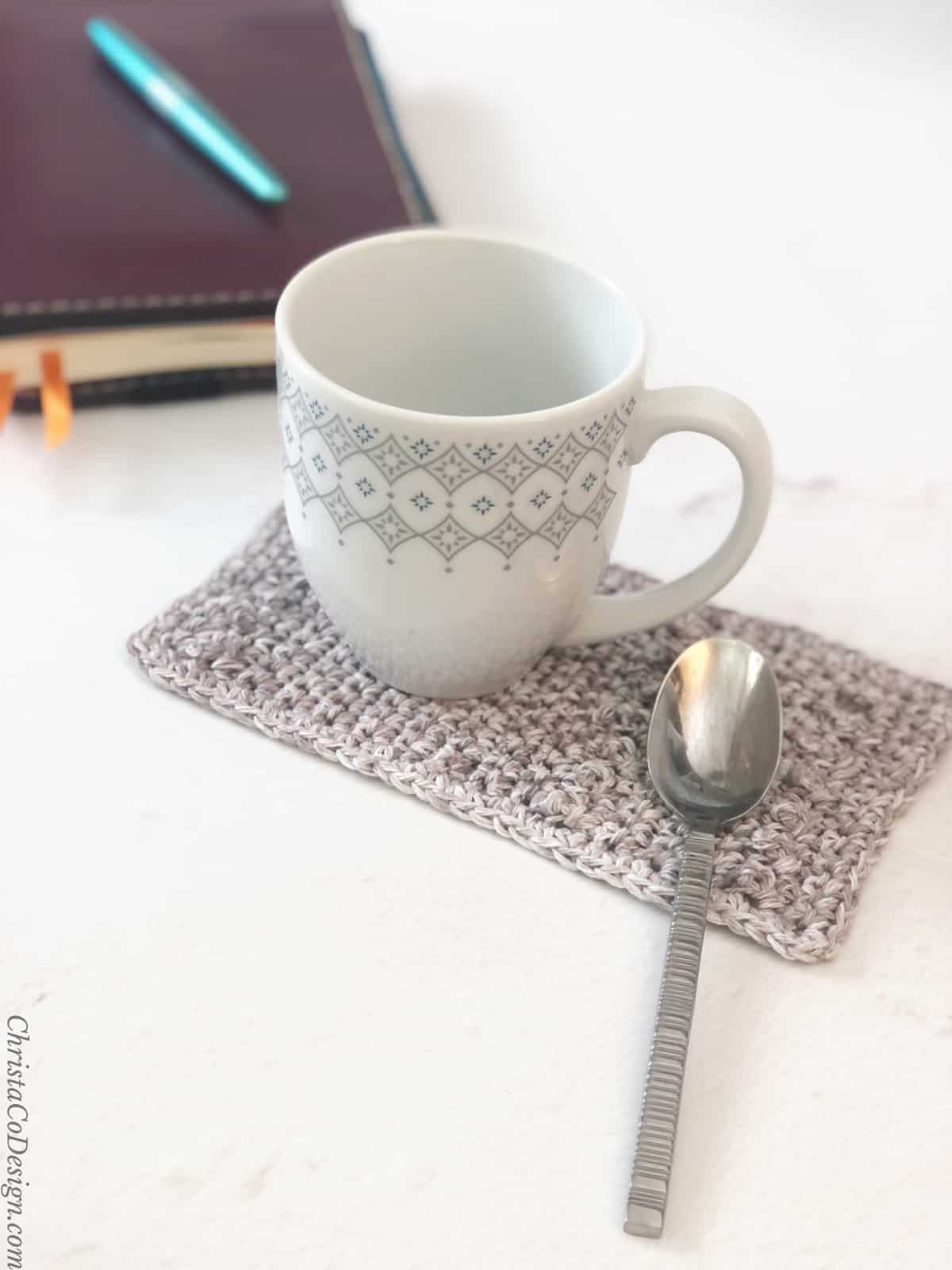 Coffee mug on grey crochet mug rug with spoon area and notebook on table.