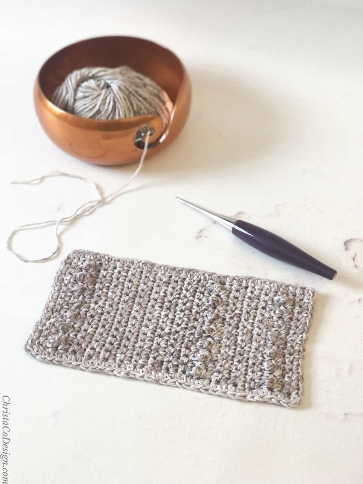 Grey mug rug crochet pattern with hook and yarn in copper bowl on table.