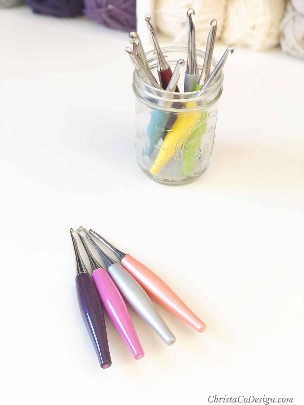 Four colorful ergonomic crochet hooks on table with more in jar behind them.