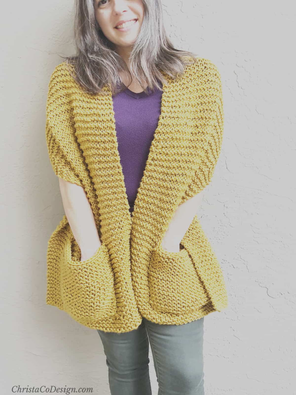 Woman with hands in pockets of mustard knit shawl.