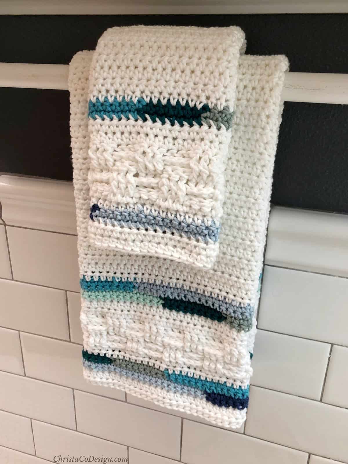 Crochet hand towel and washcloth in white with blue stripes hanging from towel bar over white subway tiles.