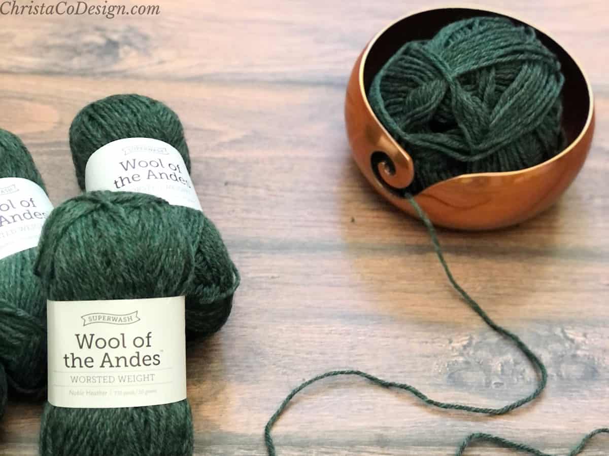 Dark green balls of yarn on wood table with one in copper yarn bowl.