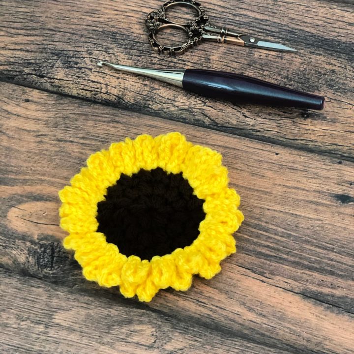 Crochet sunflower in yellow and brown on wood table with crochet hook and scissors.