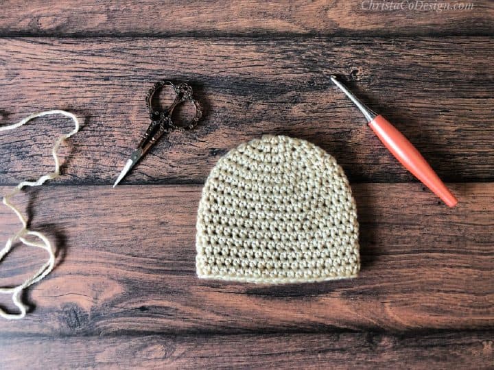 Cream colored crochet baby hat on wood table with scissors and hook.